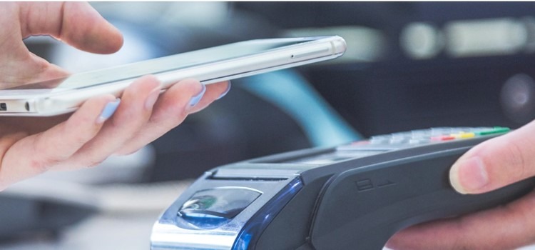 Imagem mostra a mão de uma pessoa branca aproximando um smartphone de uma máquina de cartão. O fundo da foto está desfocado. Fim da descrição.