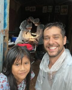 Homem sorrindo vestindo uma capa de chuva, ao lado de menina sorrindo. Ao fundo vemos dentistas em procedimento dentário em pessoa que está deitada
