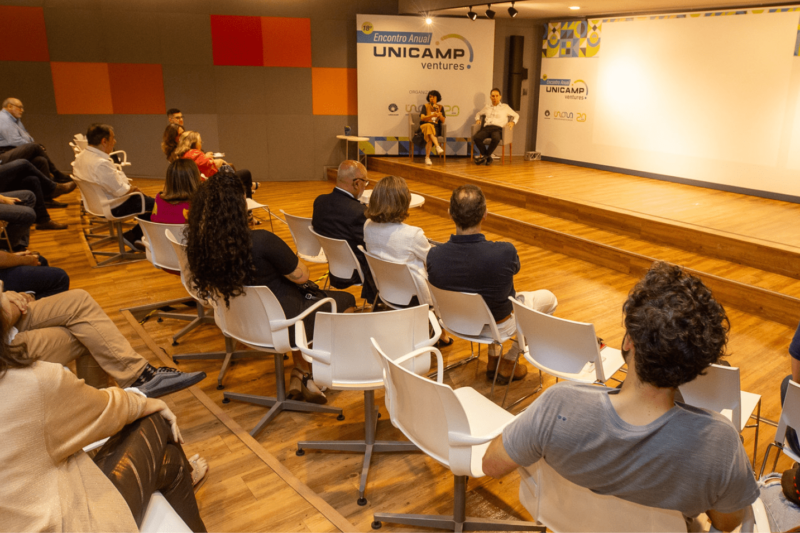 Foto colorida do auditório do Instituto Eldorado, mostram pessoas de costas e o palco onde estão um homem e uma mulher sentados no canto esquerdo da foto e uma mulher que conduz o debate no canto direito da foto em pé atrás de um púlpito. Fim da descrição.