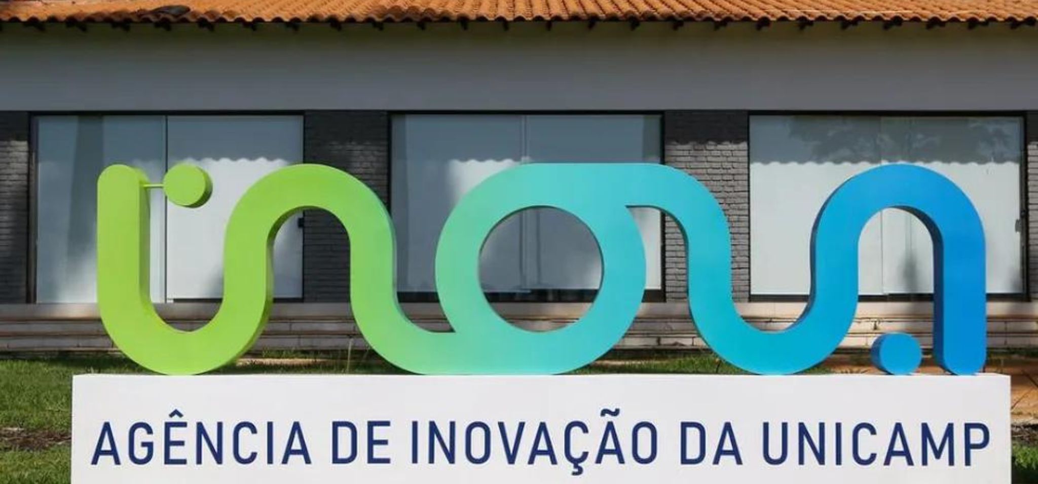 A imagem mostra uma placa da “Agência de Inovação da UNICAMP” no Brasil. A placa é composta pela palavra “Inova” em uma fonte estilizada, com as letras “I” e “n” em verde e as letras “o”, “v” e “a” em azul. A placa está sobre uma base de concreto branco com as palavras “Agência de Inovação da UNICAMP” em letras pretas. O fundo consiste em um prédio com telhado vermelho e paredes brancas.
