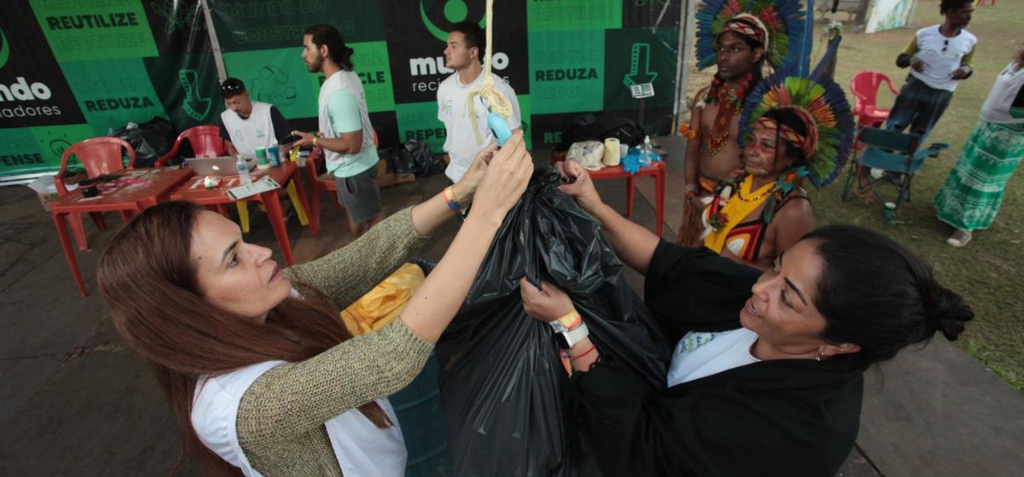 Foto de colaboradores da startup Mundo Recicladores e de participantes de um dos eventos de sustentabilidade ambiental que participam. Na imagem há duas pessoas segurando um saco plástico preto, que está pendurado em uma corda. Atrás, há pessoas, mesas e cadeiras, além de um fundo com escritos como "reutilze", "reduza". Fim da descrição.