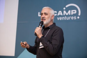 Um homem de cabelo grisalho segura um microfone e um controle remoto enquanto apresenta no palco. Ao fundo, está o logotipo do evento "Unicamp Ventures" em um painel azul. Fim da descrição.