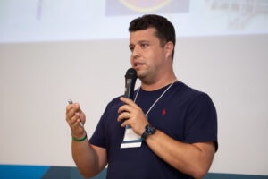 Um homem segurando um microfone e um controle remoto apresenta um tema em um ambiente de conferência. Ele usa camiseta azul escura e crachá, com um telão exibindo imagens ao fundo. Fim da descrição.