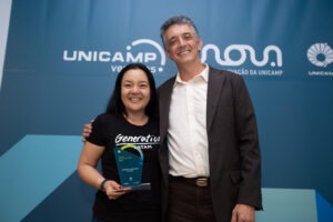 Homem entrega troféu a uma moça de camiseta preta no palco de um auditório. Ao fundo, está o logotipo do evento "Unicamp Ventures" em um painel azul. Fim da descrição.