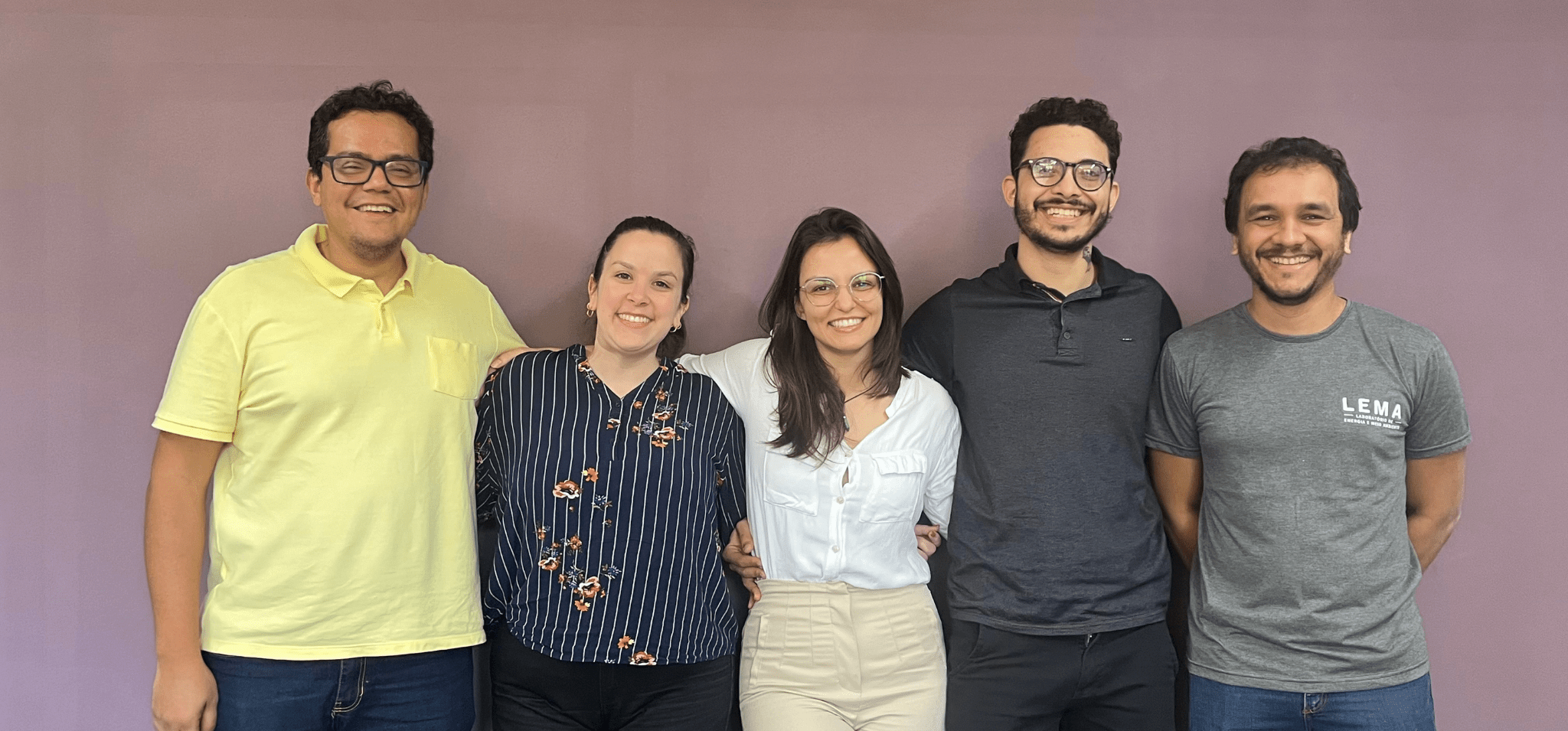 Foto de cinco pessoas adultas sorridentes posando juntos em frente a uma parede roxa. Eles estão em um ambiente interno. À esquerda, um homem usando uma camisa amarela está sorrindo e com o braço ao redor de uma mulher que usa uma camisa listrada de azul e com detalhes florais. No centro, uma mulher com uma blusa branca e óculos está sorrindo com o braço em volta de um homem de barba e óculos, que está usando uma camisa polo escura. À direita, há outro homem sorrindo, usando uma camiseta cinza com o logo "LEMA". Todos estão próximos uns dos outros e pousando para a foto. Fim de descrição.