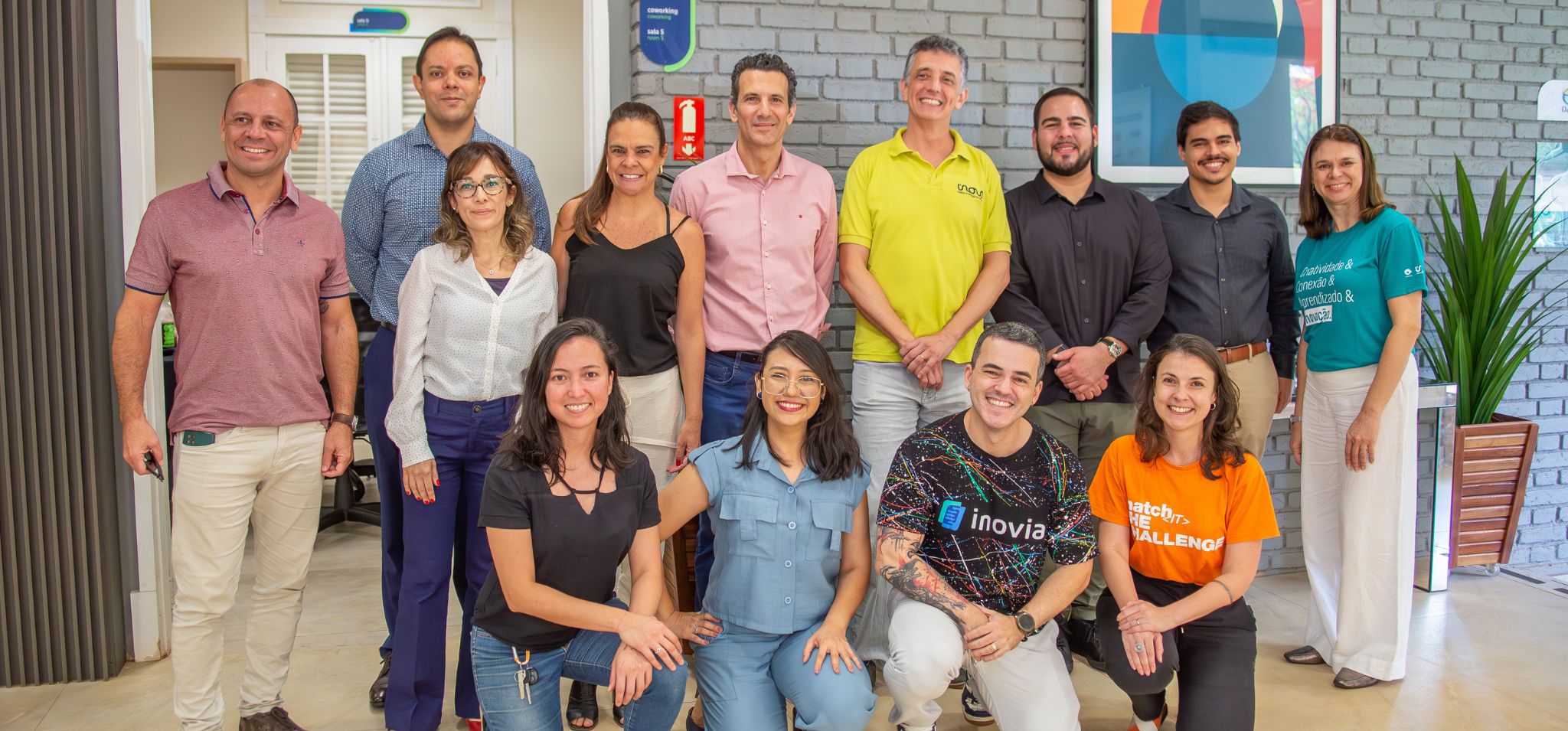 Foto posada de participantes do eMerge Americas e de colaboradores da Inova Unicamp. A imagem com treze pessoas que estão em pé ou agachadas, lado a lado. Elas estão olhando para a câmera, em ambiente interno com fundo de parede cinza e uma porta branca com vidros que dá acesso a outro local. Fim da descrição.