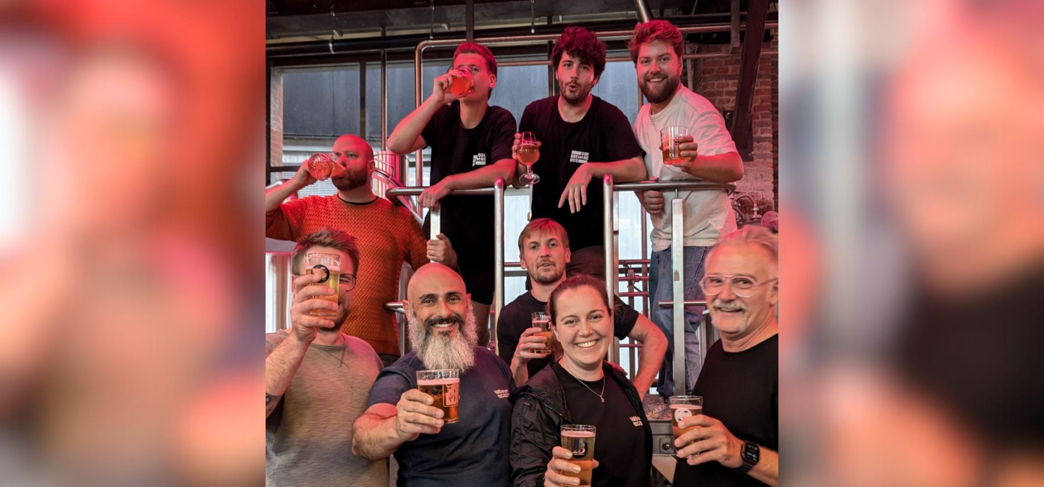 Foto colorida em espaço interno de cervejaria mostra nove pessoas sorindo e brindando com copos de cerveja. Fim da descrição.