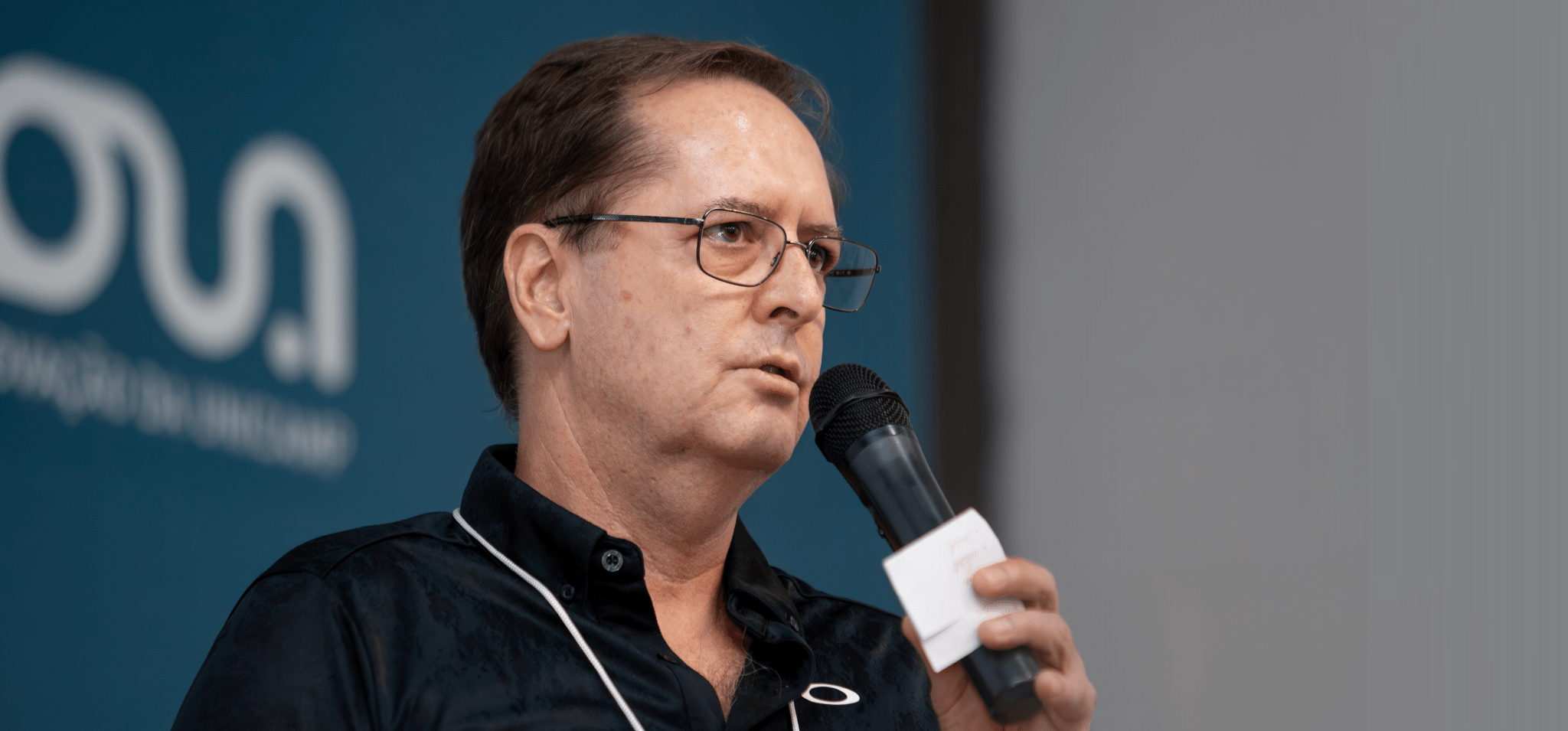 Fotografia colorida de um homem com uma microfone na mão direita. Ele está no palco fazendo sua apresentação. O fundo da fotografia está desfocado.