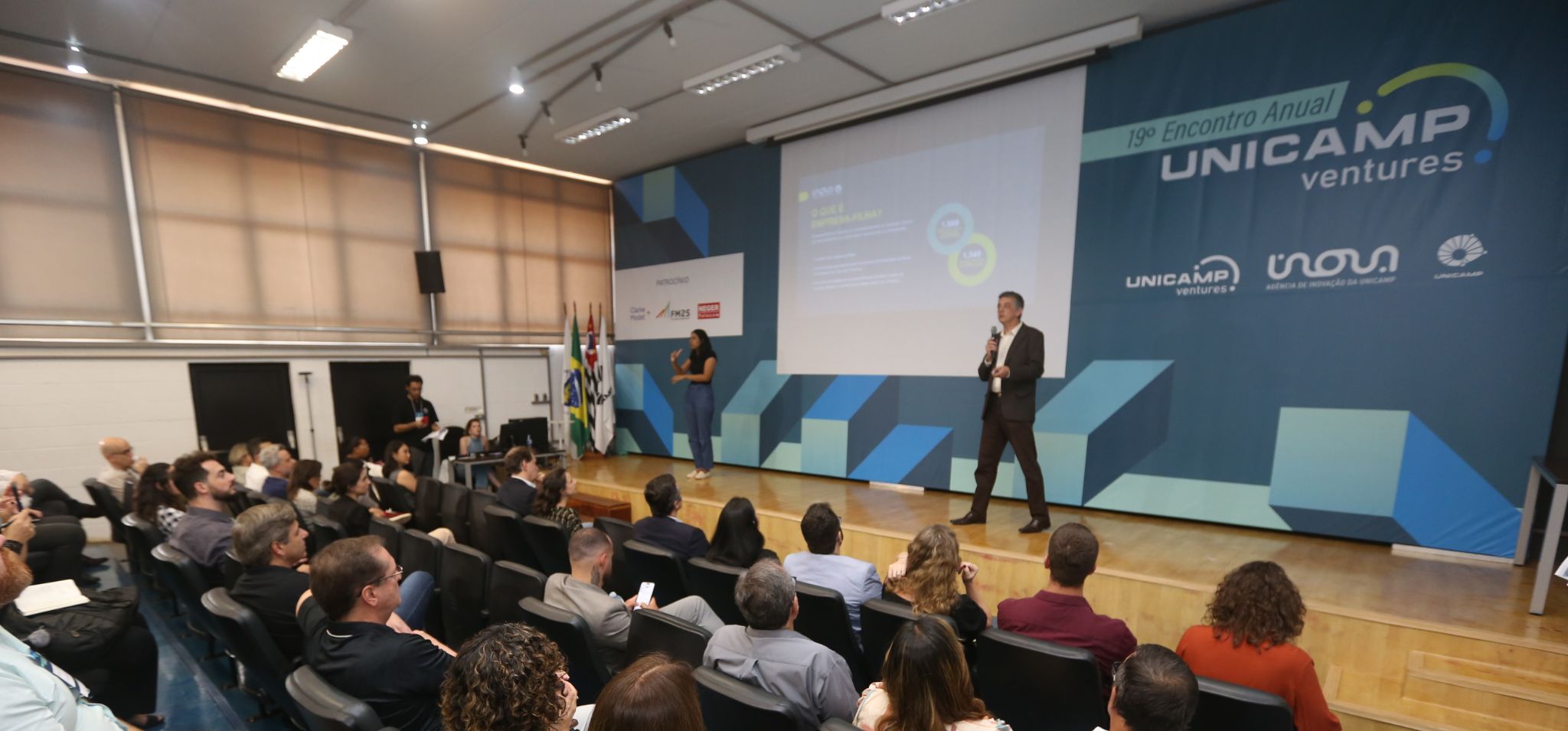 Foto colorida do professor Renato Lopes, diretor-executivo da Inova Unicamp, um homem branco vestido de terno preto e camisa branca, fazendo apresentação de slides em palco de auditório da FECFAU. Do lado esquerdo do palco há uma mulher fazendo a transcrição em libras da apresentação. Fim da descrição.