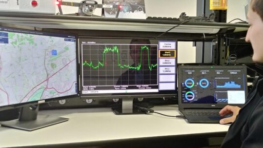 Um homem analisa dados técnicos em um laptop com gráficos, complementado por um monitor de espectro de frequência e outro com um mapa digital. A presença de um drone ao fundo sugere uma aplicação prática de monitoramento ou análise de sinais. Fim da descrição.