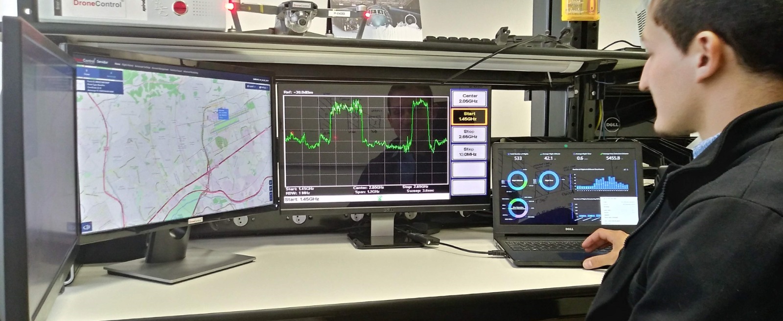Um homem analisa dados técnicos em um laptop com gráficos, complementado por um monitor de espectro de frequência e outro com um mapa digital. A presença de um drone ao fundo sugere uma aplicação prática de monitoramento ou análise de sinais. Fim da descrição.