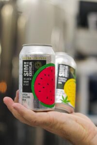 Uma mão de pele clara segura delicadamente duas latas de alumínio de cerveja sem álcool da marca "SIM! Cerveja". A lata em foco, mais próxima da câmera, apresenta um rótulo preto com o nome da marca em destaque na vertical, seguido da descrição "MELANCIA SOUR N' SALT" e um desenho estilizado de uma fatia de melancia com a casca verde e a polpa vermelha com sementes pretas. Acima, em letras menores, lê-se "EDIÇÃO LIMITADA". A segunda lata, parcialmente visível ao fundo, possui um rótulo com um design que sugere limão ou outra fruta cítrica. A mão que segura as latas está em primeiro plano, com pelos visíveis, e o fundo da imagem está desfocado, com tons claros e equipamentos industriais ou de produção levemente discerníveis. A cena transmite a apresentação e o cuidado com o produto da Empresa-filha da Unicamp, a premiada cerveja sem álcool sabor melancia.