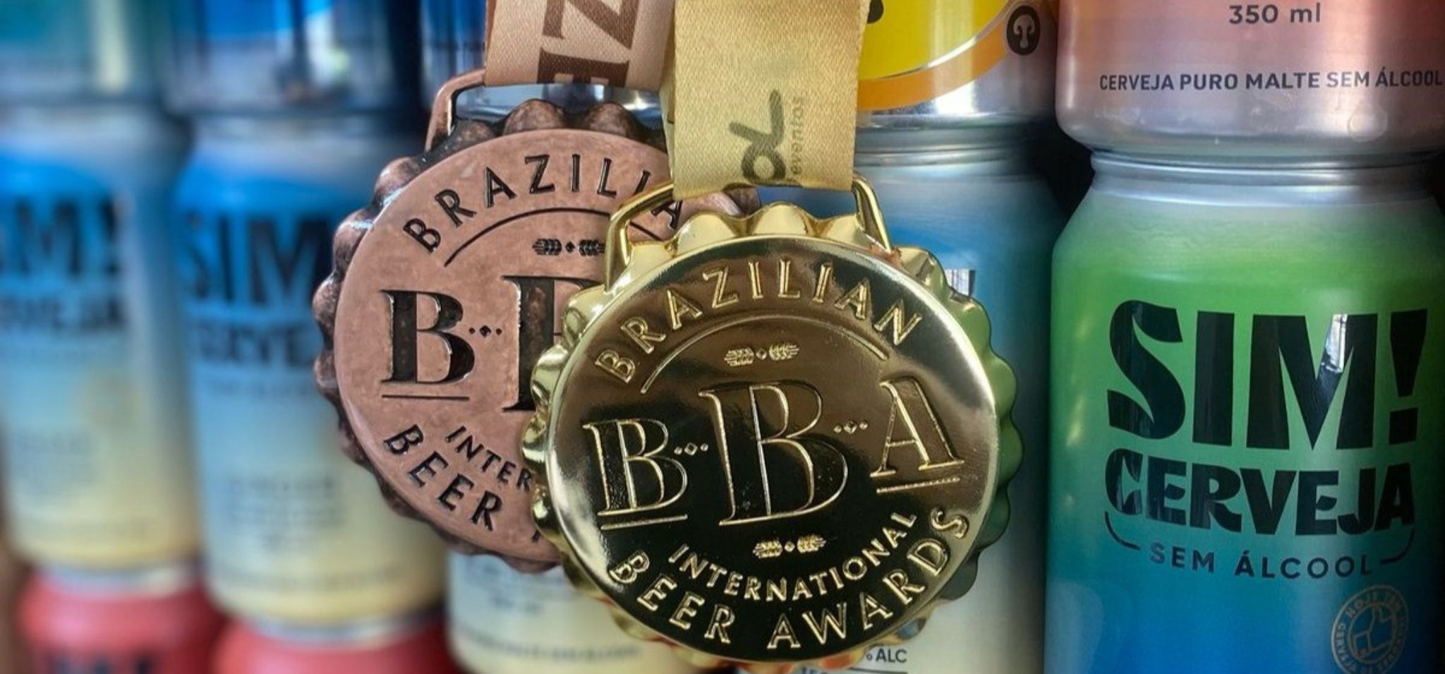A imagem exibe em primeiro plano duas medalhas de premiação da "Brazilian Beer Awards International Beer Awards". A medalha da frente é dourada e brilhante, com a inscrição "BRAZILIAN BBA INTERNATIONAL BEER AWARDS" em relevo, além das letras "BBA" em destaque no centro. A medalha de trás é de bronze, com design similar e a inscrição "BRAZILIAN BBA INTERNATIONAL BEER AWARDS" visível, embora parcialmente encoberta pela medalha dourada. Ambas as medalhas estão penduradas por fitas, sendo a da frente de cor bege e a de trás com tons terrosos. Ao fundo, desfocados, aparecem latas de cerveja da marca "SIM! Cerveja SEM álcool", alinhadas verticalmente, indicando a conquista da Empresa-filha da Unicamp é premiada na World Beer Cup 2025 com seus produtos. Empresa-filha da Unicamp é premiada na World Beer Cup 2025.