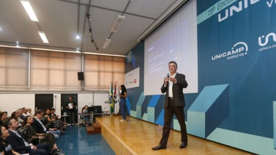 Foto de um evento da Inova Unicamp/Unicamp Ventures: um homem de meia-idade, vestindo terno escuro e camisa branca, está no palco principal de um auditório, gesticulando enquanto fala ao microfone. Atrás dele, um grande telão projeta a logo 'UNICAMP Ventures' e 'Inova Unicamp'. Uma mulher jovem, de blusa preta e calça escura, também está no palco, um pouco mais ao fundo, sendo a intérprete de libras. À esquerda, a plateia atenta é composta por diversas pessoas sentadas. A cena reflete um ambiente de palestras ou conferências sobre empreendedorismo e inovação na Unicamp. Chamada aberta: cadastre e atualize sua empresa como filha da Unicamp. Empresas-filhas da Unicamp.