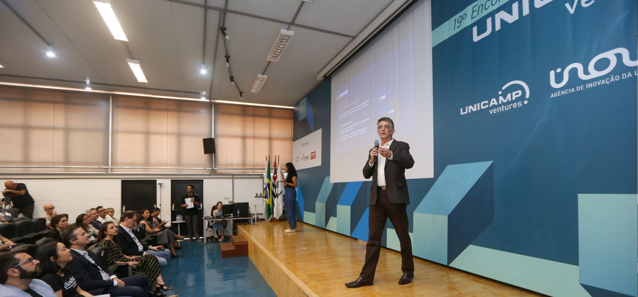 Foto de um evento da Inova Unicamp/Unicamp Ventures: um homem de meia-idade, vestindo terno escuro e camisa branca, está no palco principal de um auditório, gesticulando enquanto fala ao microfone. Atrás dele, um grande telão projeta a logo 'UNICAMP Ventures' e 'Inova Unicamp'. Uma mulher jovem, de blusa preta e calça escura, também está no palco, um pouco mais ao fundo, sendo a intérprete de libras. À esquerda, a plateia atenta é composta por diversas pessoas sentadas. A cena reflete um ambiente de palestras ou conferências sobre empreendedorismo e inovação na Unicamp. Chamada aberta: cadastre e atualize sua empresa como filha da Unicamp. Empresas-filhas da Unicamp.
