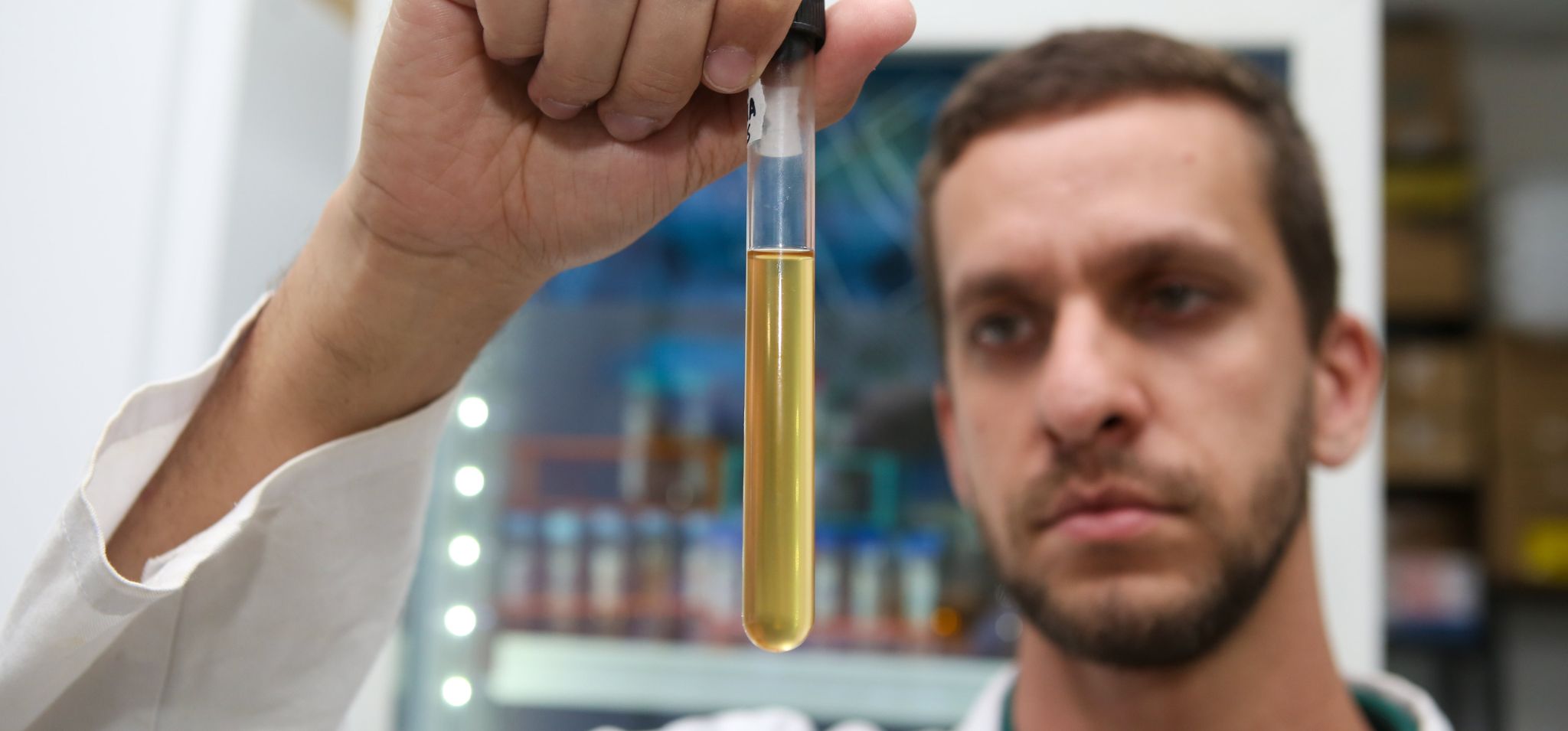 Homem de jaleco branco, em um laboratório, segura um tubo de ensaio de vidro na vertical. O tubo contém um líquido amarelo claro e transparente, e o homem o observa com atenção. Ao fundo, desfocado, percebe-se equipamentos e outros recipientes de um ambiente de pesquisa científica. Fim da descrição.