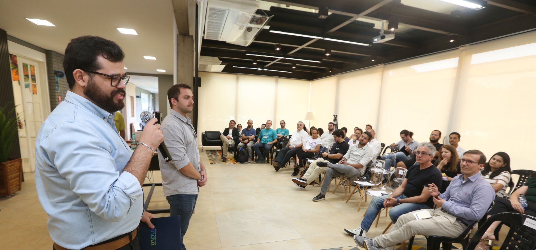 Texto alternativo: Dois homens em pé, à esquerda da imagem, conduzem uma apresentação em um ambiente corporativo. Um deles segura um microfone e uma pasta com o logotipo da Inova Unicamp. À direita, várias pessoas estão sentadas em cadeiras, prestando atenção à fala. Fim da descrição.