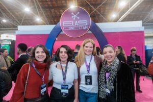 Quatro mulheres sorrindo, posam juntas em frente a um estande do evento "Elas Conectam", identificado por um grande arco rosa com um letreiro circular roxo. Todas as mulheres usam crachás de identificação do evento. O ambiente parece ser de uma feira ou congresso.