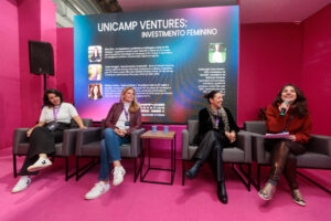 Quatro mulheres participam de um painel de discussão sentadas em poltronas no palco. Uma mulher na ponta direita está falando ao microfone. O painel luminoso ao fundo exibe o título "UNICAMP VENTURES: INVESTIMENTO FEMININO" e as fotos e descrições das palestrantes.