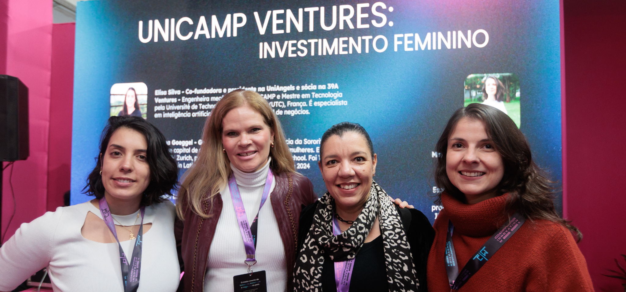 Quatro mulheres sorriem para a câmera em frente a um painel com o texto "UNICAMP VENTURES: INVESTIMENTO FEMININO". Elas estão usando crachás de evento, e o painel ao fundo contém pequenos retratos e informações biográficas.