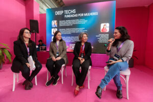 Quatro mulheres sentadas em cadeiras num palco, interagindo durante um painel de discussão. Ao fundo, um telão exibe o título "DEEP TECHS FUNDADAS POR MULHERES" e perfis das palestrantes. Uma das mulheres segura um microfone. O cenário é em tons de rosa e roxo.