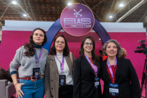 Quatro mulheres posam sorrindo em frente a um grande arco decorado com um letreiro circular que diz "ELAS CONECTAM" e tem um símbolo de rede ou conexão no centro. Todas usam crachás de evento.