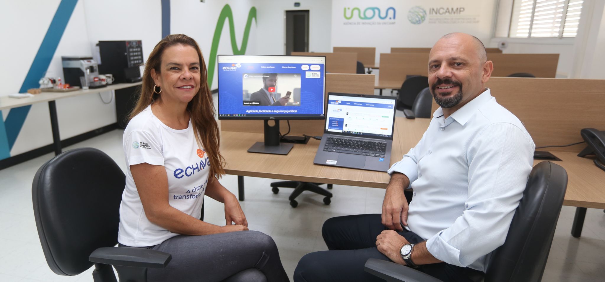Duas pessoas estão sentadas em cadeiras giratórias em frente a uma mesa com um monitor e um notebook em uma sala de escritório. À esquerda, uma mulher com cabelo castanho comprido veste uma camiseta branca com o logotipo da eChave e calça jeans escura. À direita, um homem calvo com barba, usando camisa social branca e calça escura, sorri para a câmera. A tela do monitor e do notebook exibe a interface do site do projeto. Ao fundo, há estações de trabalho vazias e logotipos nas paredes indicando 'Inova Unicamp' e 'Incamp’. Fim da descrição.