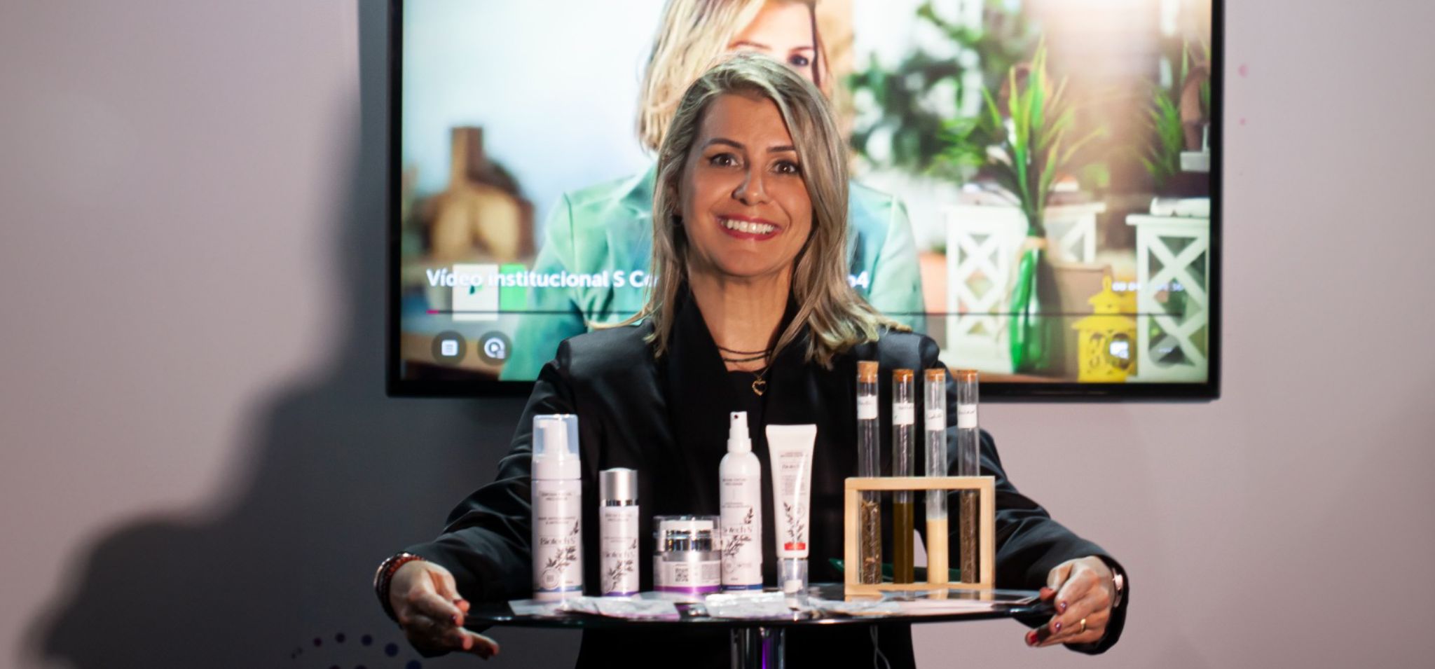 A imagem mostra uma mulher de meia-idade, cabelos loiros lisos na altura dos ombros, e um sorriso acolhedor. Ela está usando um blazer preto sobre uma blusa de cor clara e segura com as duas mãos uma bandeja transparente, que contém diversos produtos cosméticos e alguns tubos de ensaio. À esquerda da bandeja, há cinco embalagens de produtos cosméticos em tons claros (branco, cinza e lilás), com designs elegantes e rótulos que parecem conter informações sobre a marca. À direita, sobre um pequeno suporte de madeira, estão dois tubos de ensaio com líquidos amarelados e alguns outros itens de laboratório. A mulher está posicionada atrás de uma mesa, e ao fundo, há uma tela grande exibindo um vídeo institucional. O ambiente parece ser um estúdio ou sala de apresentação, com iluminação focada na pessoa e nos produtos. A parte inferior da imagem revela um pequeno detalhe de um logotipo ou design gráfico em tons de azul.