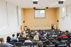 Uma visão ampla de uma sala de palestras moderna, onde um orador de terno escuro está em pé no palco à direita, apresentando a um público sentado em cadeiras de auditório. No centro do palco, um grande projetor exibe um slide com o texto "Quem somos" e uma imagem de uma paisagem verde. A sala tem paredes de madeira clara na frente e paredes brancas nas laterais, com painéis acústicos pretos e luzes no teto. Há também algumas lixeiras coloridas no canto esquerdo da sala. Empresas-filhas da Unicamp impulsionam inovação verde com relevância global e nacional.