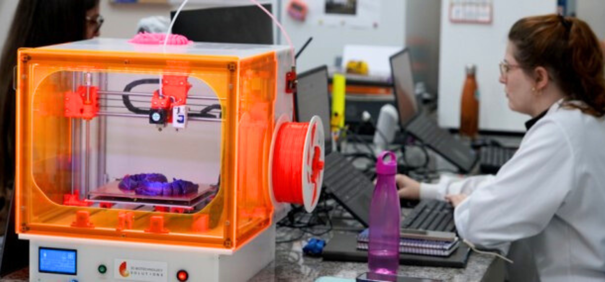 A imagem mostra um laboratório ou espaço de trabalho com uma impressora 3D laranja em primeiro plano, que está em operação imprimindo um objeto roxo. Há um rolo de filamento vermelho próximo à impressora. À direita, uma mulher de jaleco branco e óculos está sentada em frente a um laptop e um monitor, digitando. Ao fundo, outra pessoa pode ser vista sentada à esquerda. Fim da descrição.