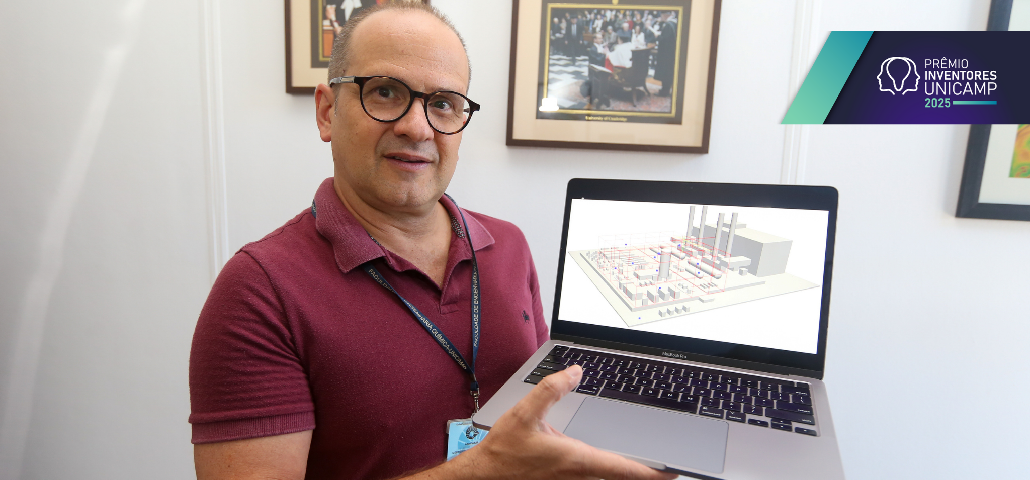 A imagem mostra um homem de óculos e camisa polo bordô segurando um notebook aberto com uma das mãos e apontando para a tela com a outra. Na tela do computador, é exibido um modelo tridimensional de uma planta industrial ou instalação técnica, com estruturas retangulares e tubulações visíveis. O homem está em um local que remete a um ambiente acadêmico. Fim da descrição.