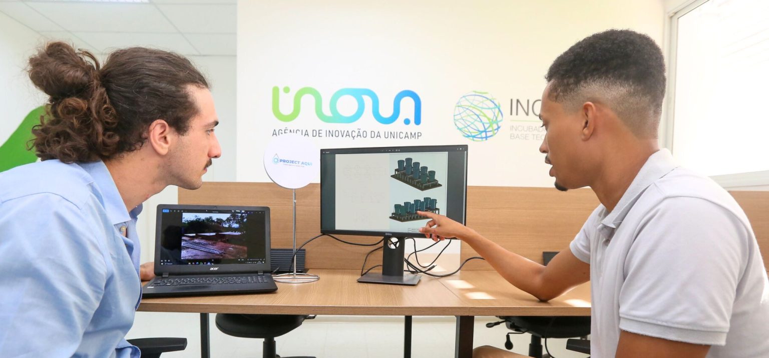 Dois homens estão sentados frente a frente em uma sala de reuniões, observando dois monitores. O homem à direita aponta para a tela de um monitor que exibe esquemas técnicos e projeções tridimensionais de estruturas, enquanto o homem à esquerda observa atentamente e segura um notebook com uma imagem de paisagem natural. Ao fundo, há um banner com os logos da Inova Unicamp e da Incubadora de Base Tecnológica. Fim da descrição.