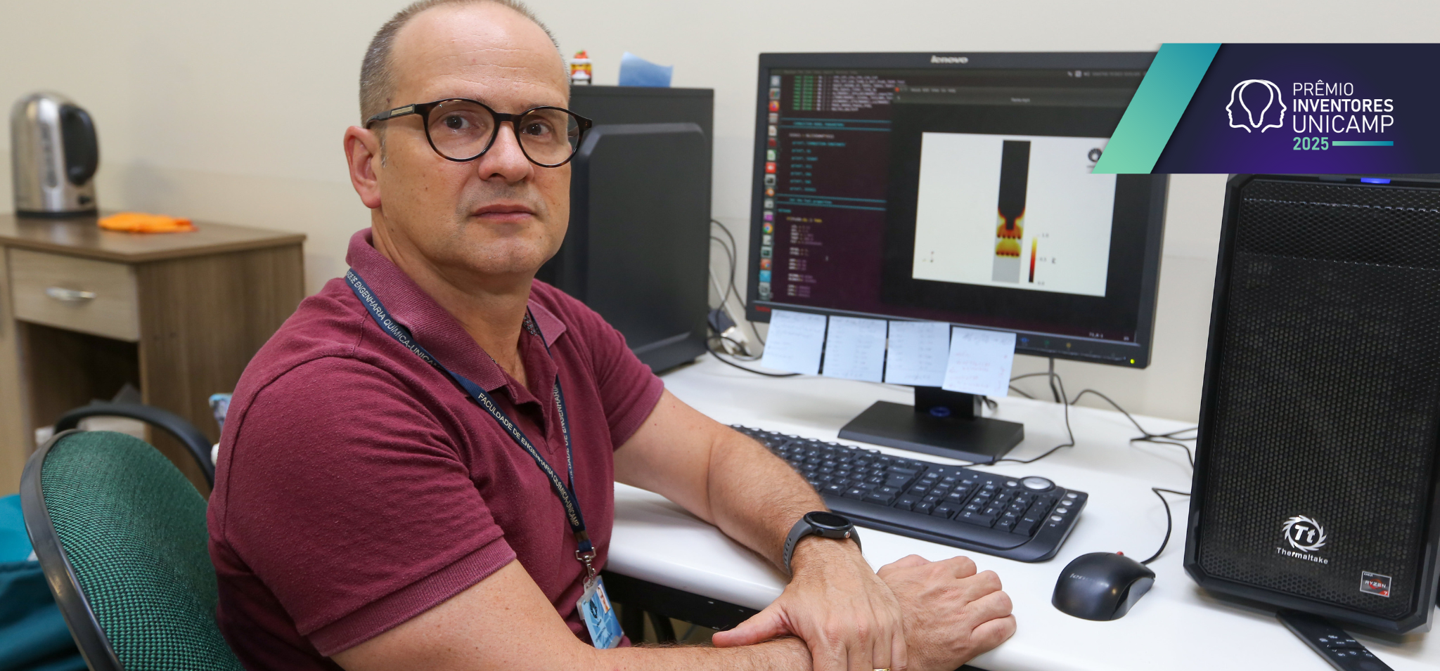 A imagem mostra um homem de óculos e camisa polo bordô sentado em uma cadeira de escritório, posicionado em frente a uma mesa de trabalho e olhando diretamente para a câmera. Sobre a mesa, à sua frente, há uma tela de computador exibindo um diagrama técnico. Ao lado da mesa, veem-se outros objetos de escritório. O ambiente geral sugere um escritório acadêmico. Fim da descrição.