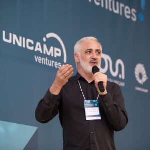 Juliano Prado, gerente de Propriedade Intelectual da Inova Unicamp, fala em um microfone com as mãos gesticulando, em um evento da Unicamp Ventures. O logo da Unicamp Ventures é visível ao fundo.
