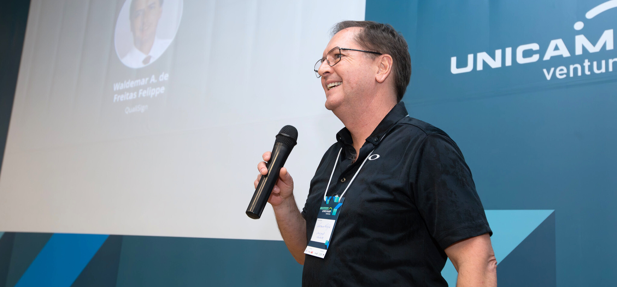 Waldemar Felippe, presidente-executivo do conselho do Unicamp Ventures, sorri enquanto segura um microfone e discursa em um evento, com o logo 'Unicamp Ventures' ao fundo. Comunidade Unicamp Ventures Empresa-filha.