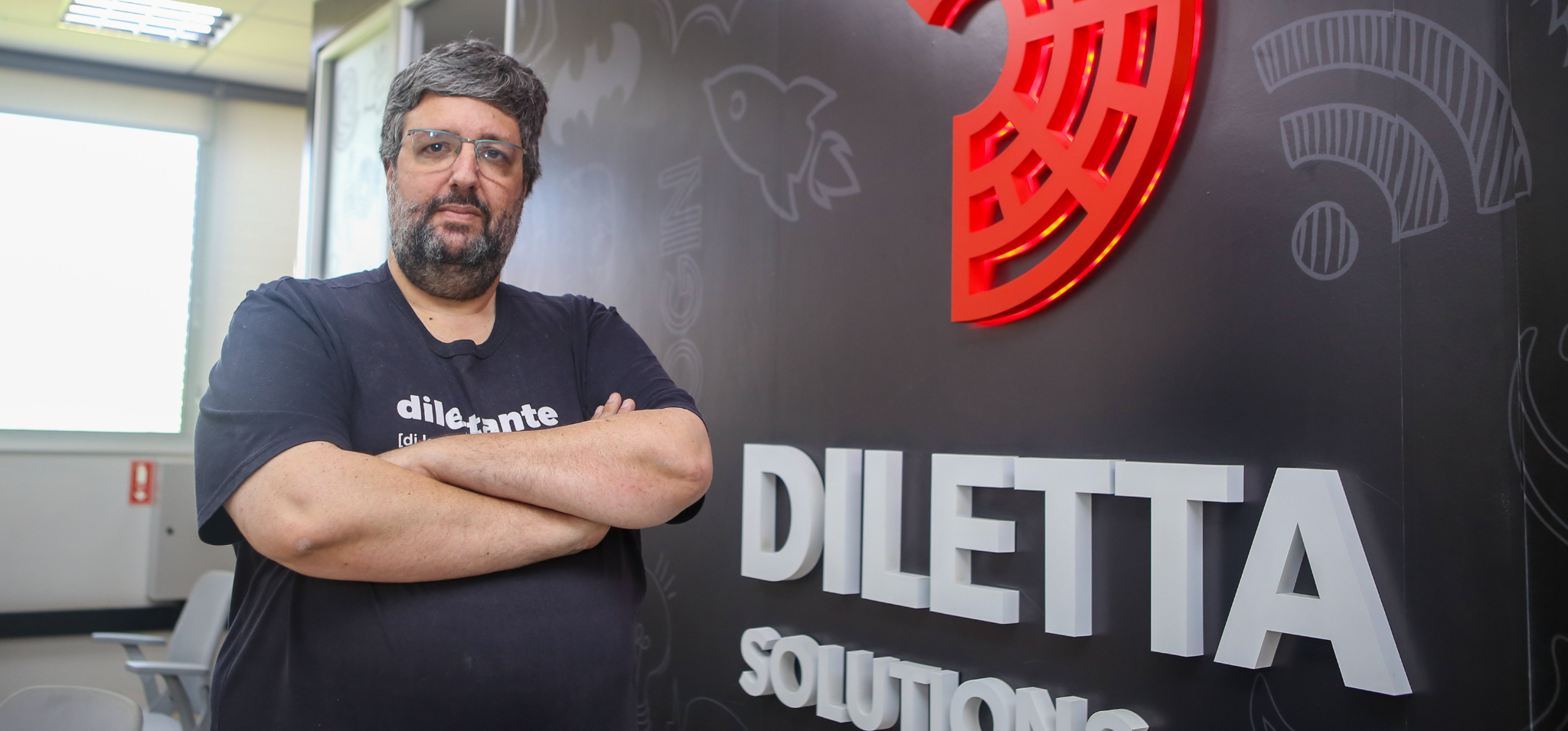 Um homem barbudo com óculos e uma camiseta preta com a palavra "Dilettante" estampada, posa com os braços cruzados na frente de uma parede preta com o logotipo vermelho e branco da "Diletta Solutions". Há ícones de tecnologia e inovação desenhados em branco na parede. Fim da descrição.