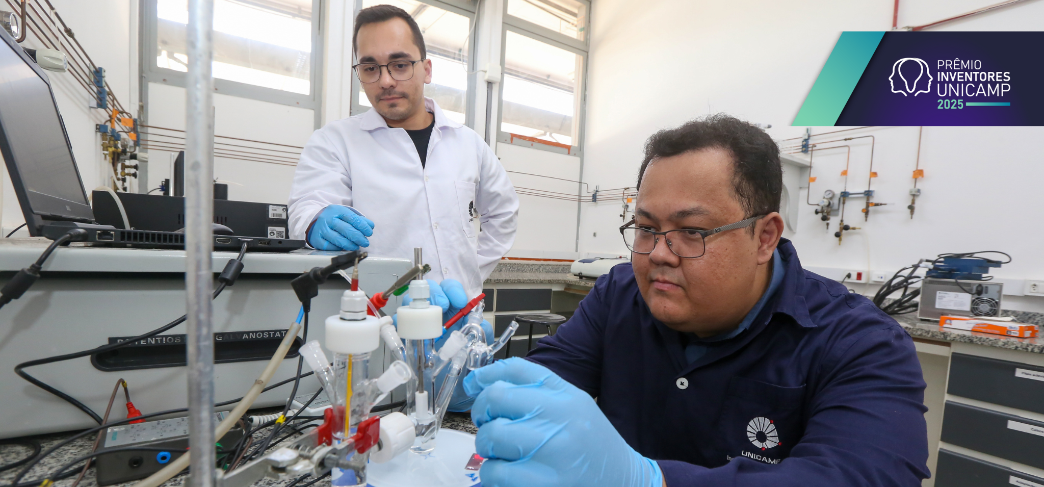 Dois pesquisadores em um laboratório, manipulando um experimento eletroquímico. À direita, um homem de cabelos curtos e óculos, vestindo um jaleco azul marinho com o logo da Unicamp e luvas azuis, está sentado e manuseia cuidadosamente um equipamento com vidrarias e eletrodos. À esquerda, um homem de cabelos curtos, barba curta, vestindo um jaleco branco e luvas azuis, está em pé, observando atentamente o experimento. O ambiente é um laboratório com bancadas de granito, equipamentos científicos, um computador e conexões elétricas e hidráulicas aparentes nas paredes. Há janelas ao fundo permitindo a entrada de luz natural. As cores predominantes são branco, cinza e azul. Fim de descrição.