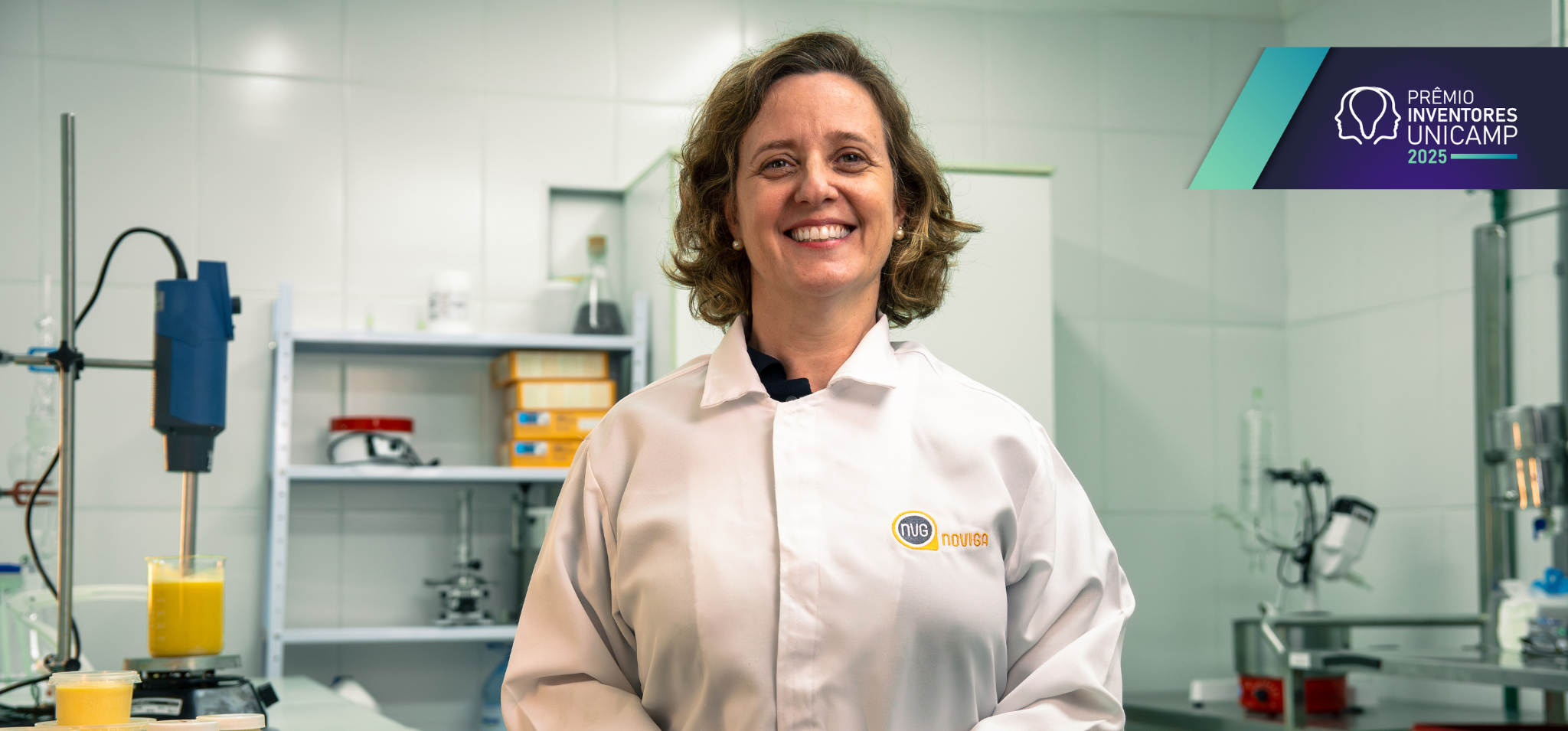 Retrato de corpo inteiro de uma mulher com cabelos castanhos claros na altura dos ombros, sorrindo para a câmera. Ela veste um jaleco branco de laboratório com o logotipo "NOVIGA" e está em um laboratório. Ao fundo, pode-se ver equipamentos de laboratório, incluindo um misturador e um béquer com um líquido amarelo. No canto superior direito da imagem, há o logotipo "PRÊMIO INVENTORES UNICAMP 2025". Fim da descrição.