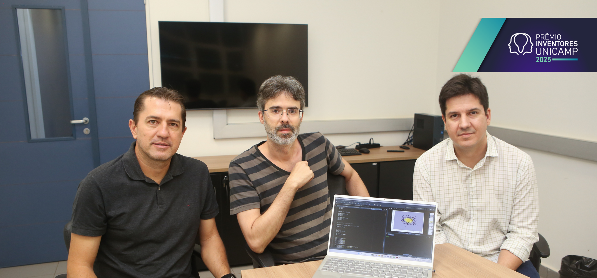 Três homens posam para uma foto em uma sala de reunião, sentados ao redor de um laptop. O homem à esquerda veste uma camisa polo preta. O homem ao centro tem barba grisalha e usa uma camisa listrada. O homem à direita usa uma camisa xadrez branca. O laptop à frente mostra um gráfico na tela. No canto superior direito da imagem, há um logotipo com a inscrição "PRÊMIO INVENTORES UNICAMP 2025". Fim da descrição.