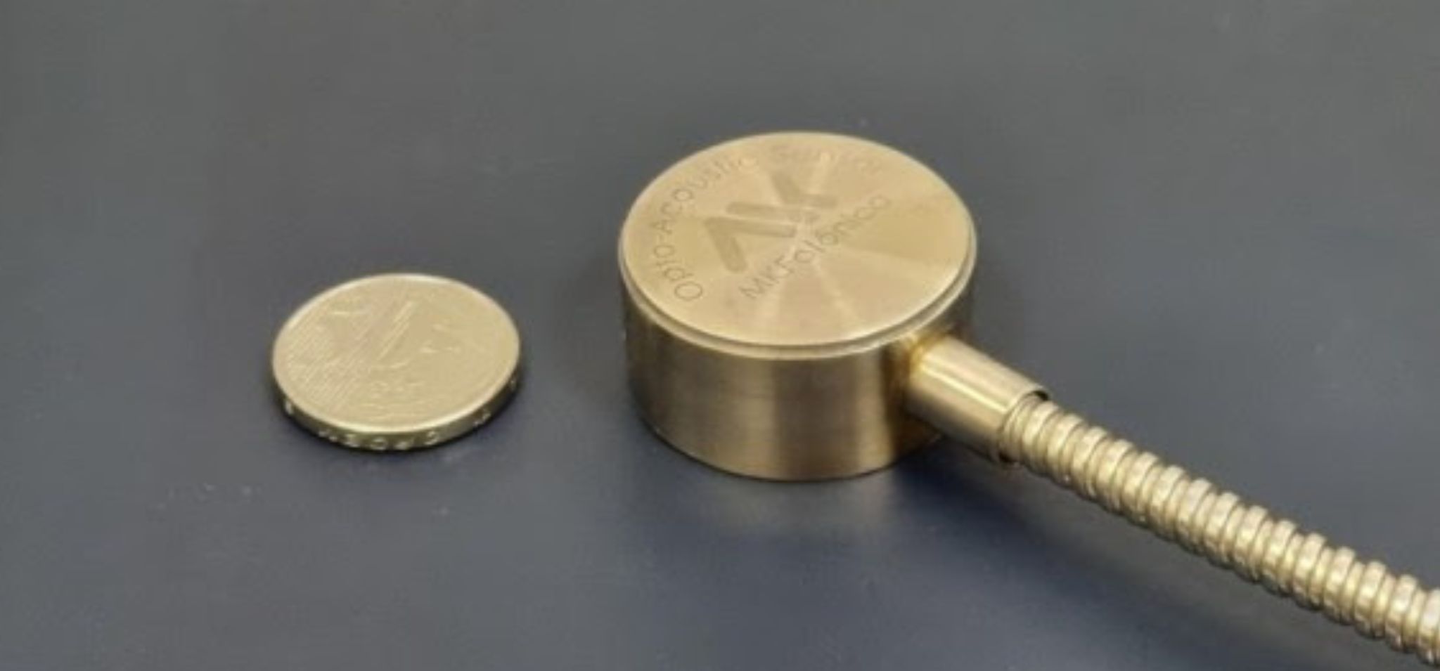 Na imagem, há uma moeda de 1 real posicionada ao lado de um dispositivo metálico cilíndrico dourado, conectado a um cabo flexível revestido, onde está escrito MK Fotônica. Fim da descrição.