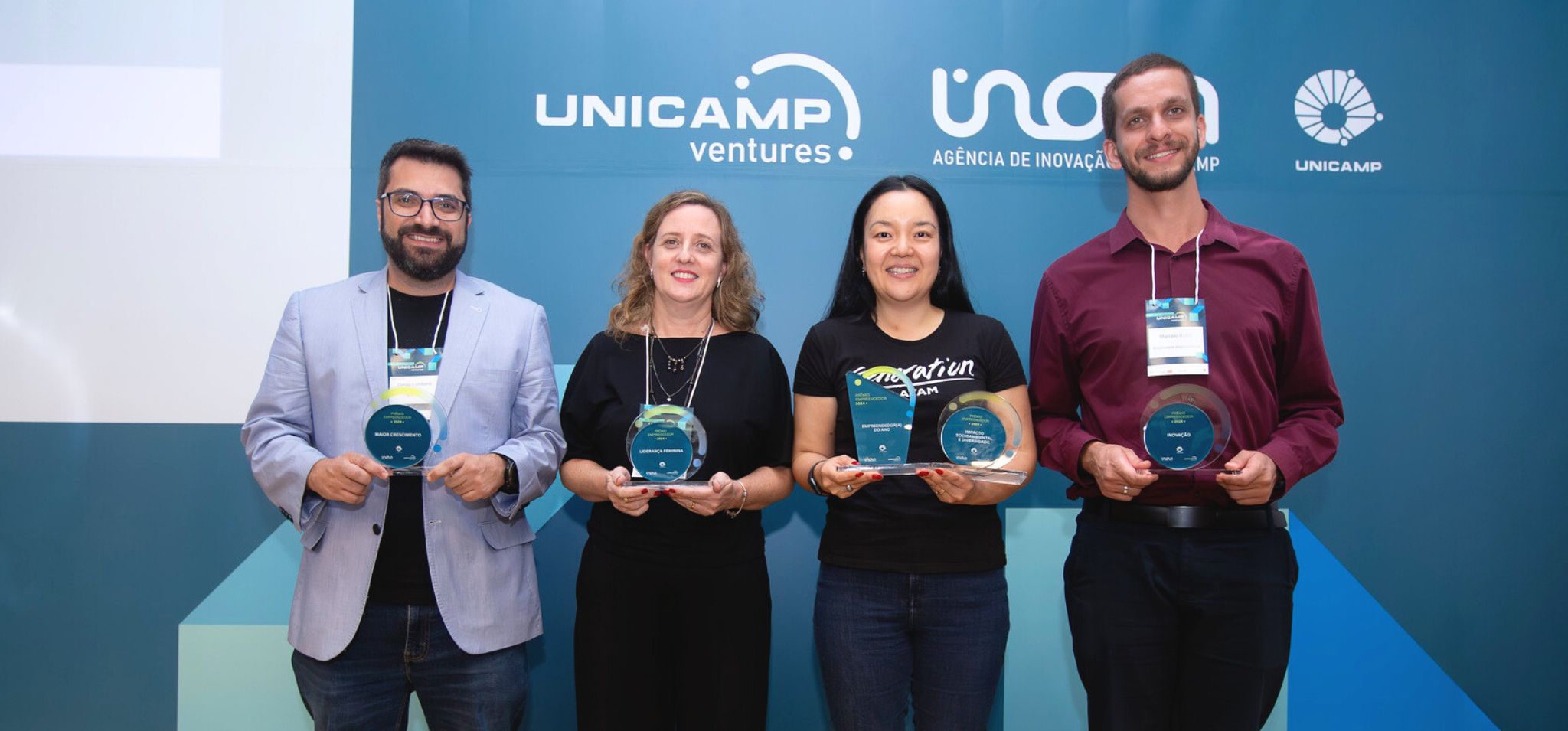 Quatro pessoas vencedoras do Prêmio Empreendedores Unicamp, duas mulheres e dois homens, estão em pé lado a lado, sorrindo para a câmera. Ao fundo, uma parede com os logotipos "UNICAMP ventures" e "Inova Agência de Inovação". Cada pessoa segura um prêmio de acrílico em formato redondo. Da esquerda para a direita: um homem com barba e óculos, vestindo um blazer claro; uma mulher com cabelo loiro na altura dos ombros, usando uma blusa preta; uma mulher com cabelo escuro e comprido, usando uma camiseta preta com a palavra "innovation" estampada; e um homem com barba rala, usando uma camisa roxa. Prêmio Empreendedores Unicamp