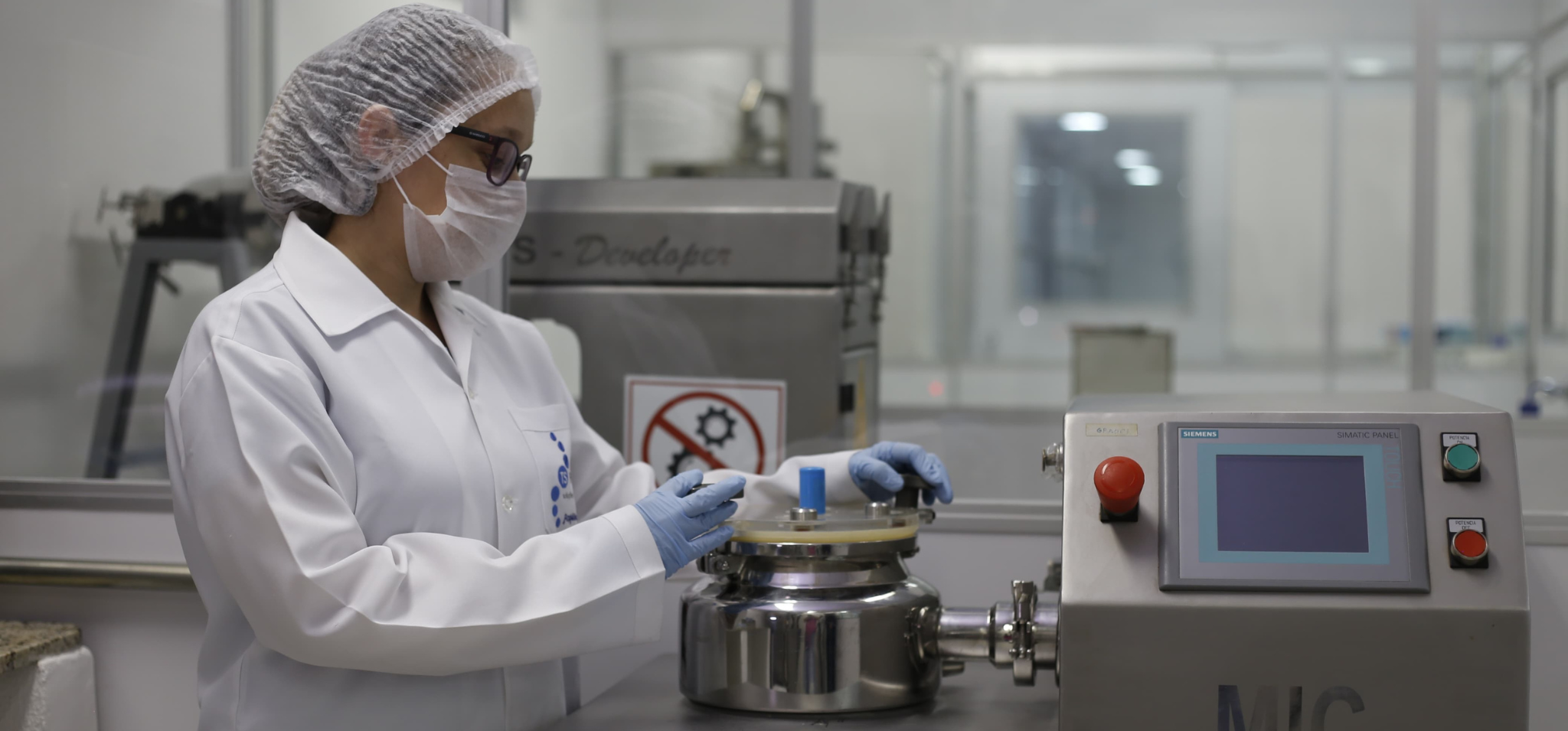 Uma foto que mostra uma mulher branca, com um jaleco azul-escuro e luvas de látex rosas. Ela está no interior de um laboratório com paredes de cor cinza e vidro, posicionada entre duas bancadas de granito, em um ambiente esterilizado e com iluminação clara. Ela está concentrada e despejando um líquido de um frasco menor para outro maior, de formato cônico, que contém um líquido vermelho. Fim da descrição.
