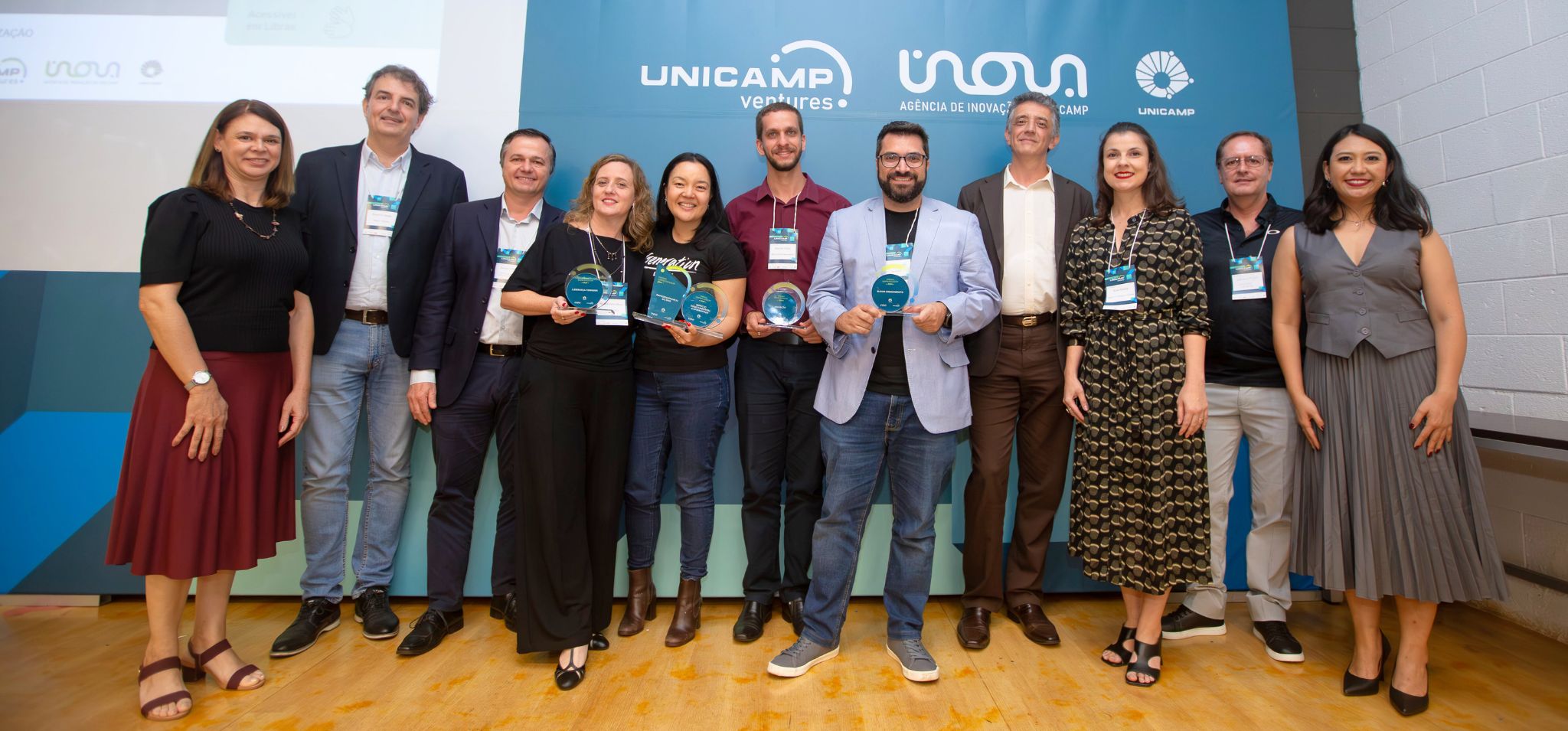 É uma foto de um grupo de dez pessoas adultas. A cena acontece em um ambiente interno (auditório ou palco) durante um evento, com um piso de madeira clara e um painel de fundo em tons de azul e branco, que exibe as logomarcas da UNICAMP Ventures e INOVA Agência de Inovação UNICAMP. A iluminação é forte e focada no grupo. O grupo de dez pessoas (seis homens e quatro mulheres) está em pé, lado a lado, sorrindo para a câmera. Sete delas seguram troféus de vidro azulados, indicando uma premiação. O grupo usa trajes casuais de negócios variados, predominando tons escuros. Fim de descrição. Eles se reuniram para o 20º encontro anual Unicamp Ventures.