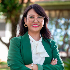 É uma foto em plano médio, com as bordas arredondadas. O assunto principal é uma mulher adulta, identificada pelo nome do arquivo como Kátia Kishi. A paisagem é em uma área externa, provavelmente um jardim, com o fundo desfocado em tons de verde e marrom. A luz é natural e suave. A ação é a mulher está em pé, sorrindo para a câmera com os braços cruzados. Ela usa um blazer verde-escuro, blusa branca com laço na gola, óculos de aro dourado e brincos de argola dourada. Prêmio Empreendedores Unicamp 2025. Fim de descrição.