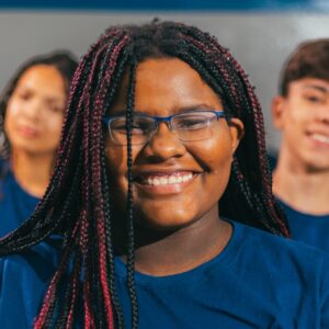 Fotografia. A imagem mostra um close-up de uma jovem negra sorrindo diretamente para a câmera. Ela tem cabelos longos com tranças escuras, algumas com mechas vermelhas, e usa óculos de armação azul. Veste uma camiseta azul lisa. A jovem está com um sorriso largo, mostrando os dentes. Atrás dela, desfocados, há dois outros jovens: um à esquerda, com pele morena e cabelos escuros, e outro à direita, de pele clara e cabelo escuro, ambos também sorrindo e vestindo camisetas azuis, complementando a cena com expressões alegres. A iluminação é suave e uniforme, focando na expressão facial da jovem em primeiro plano. Fim de descrição.