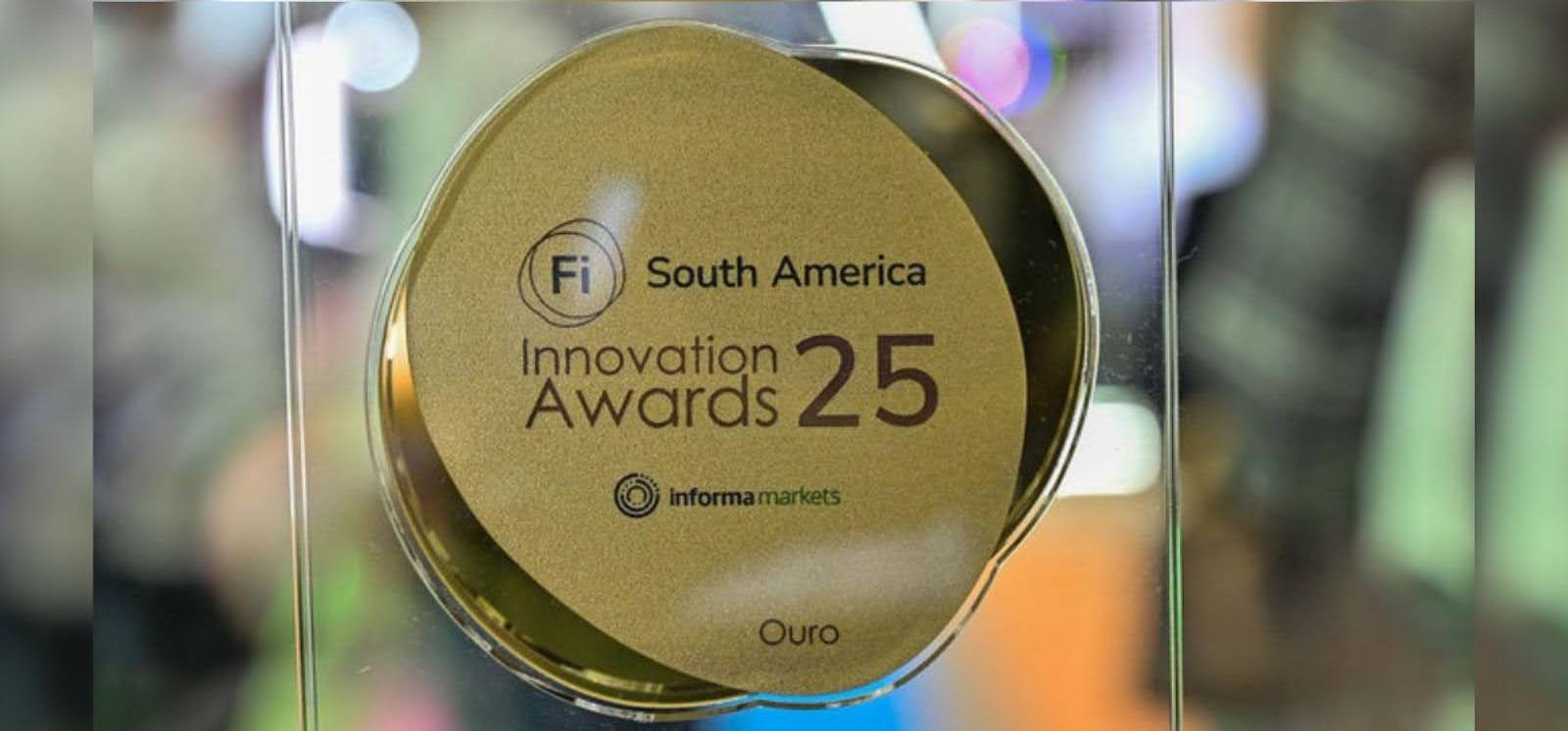 A imagem mostra uma medalha dourada e oval, com a inscrição "Fi South America Innovation Awards 25". Abaixo, em letras menores, está "informa markets" e, na parte inferior, a palavra Ouro. A medalha está exposta sobre uma superfície transparente, refletindo a luz e o ambiente ao redor. Fim da descrição.