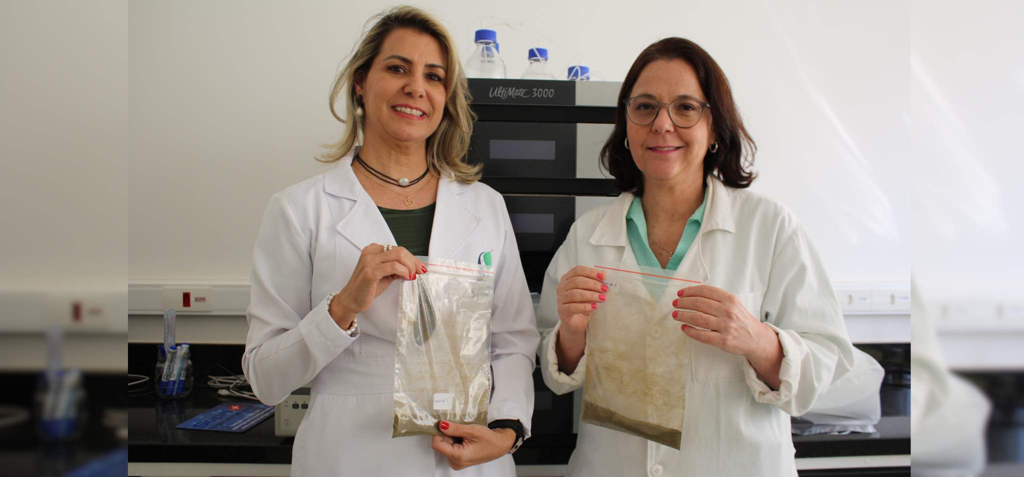 Na imagem, há duas mulheres vestindo jaleco branco e segurando sacos com resíduos de extração da planta Artemisia annua. Fim da descrição.