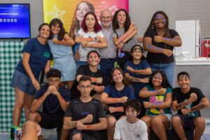 Fotografia. A imagem mostra um grupo de cerca de 15 adolescentes e dois adultos posando em frente a uma parede colorida com tons de amarelo, laranja e rosa, e com um banner que exibe fotos de jovens e a frase "Eu sou Maah". Os adolescentes, de várias etnias, estão vestidos com roupas casuais, em sua maioria camisetas e shorts ou calças jeans. Alguns têm palavras como "QUÍMICA", "ELETRÔNICA", "COTUCA" e "BENTO" escritas em seus braços com tinta facial. Um homem idoso, de pele clara, cabelos brancos e vestindo uma camisa xadrez, está no centro superior do grupo, sorrindo e com os braços cruzados. Todos os jovens estão sorrindo e muitos fazem gestos com os braços cruzados ou com os punhos cerrados. À esquerda da imagem, parte de uma tela de televisão exibe números, e à direita, uma área com uma bancada branca sugere um ambiente de cozinha ou laboratório. Há uma mistura de cores vibrantes no fundo e nos detalhes, com os tons de pele e as roupas dos participantes. Fim de descrição.