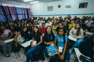 Fotografia. A imagem mostra um grupo de cerca de 30 adolescentes em uma sala de aula, dispostos em fileiras de carteiras. No centro, uma mulher adulta de pele clara, cabelos loiros, e vestindo uma roupa verde clara, está sorrindo. Ela está rodeada por jovens de diversas etnias, que também sorriem para a câmera. Alguns dos jovens estão acenando ou fazendo um "joinha" com a mão. As cores predominantes são o azul das calças jeans e os tons variados das camisetas dos estudantes. A sala de aula tem paredes brancas e uma cortina azul escura do lado esquerdo. Ao fundo, uma faixa com a inscrição "MUDANDO A HISTÓRIA" é visível. A iluminação é difusa, indicando um ambiente interno bem iluminado. Fim de descrição.