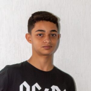 Fotografia. A imagem é um close-up de um jovem de pele clara, com cabelo curto e escuro, vestindo uma camiseta preta com uma estampa branca. Ele olha diretamente para a câmera com uma expressão séria. O fundo é uma parede branca com textura. A iluminação é suave e o foco está no rosto do jovem. Fim de descrição.